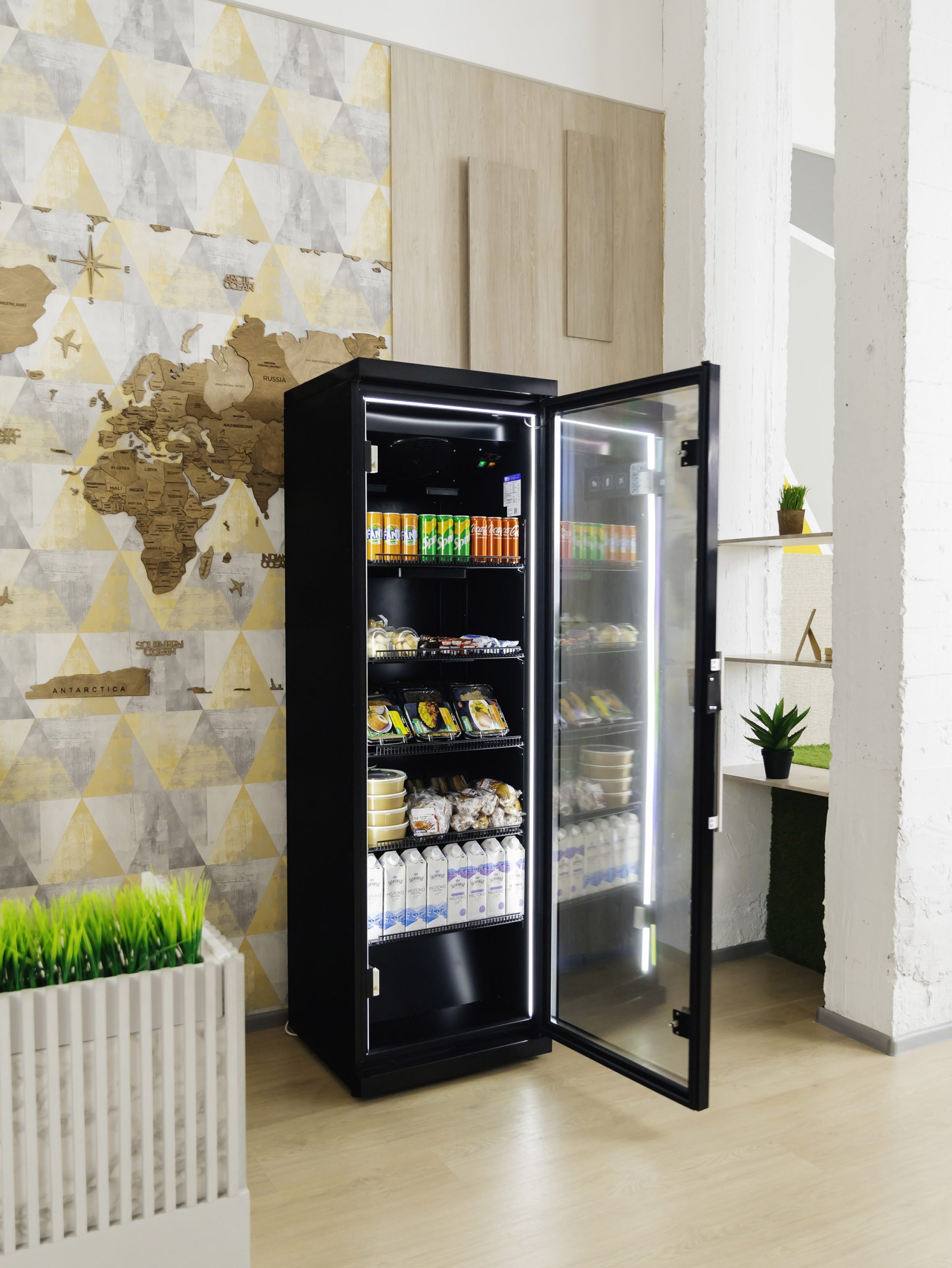 Black cabinet with the glass door open in a room with a decorative wall panel and plants, drinks and snacks on the shelves